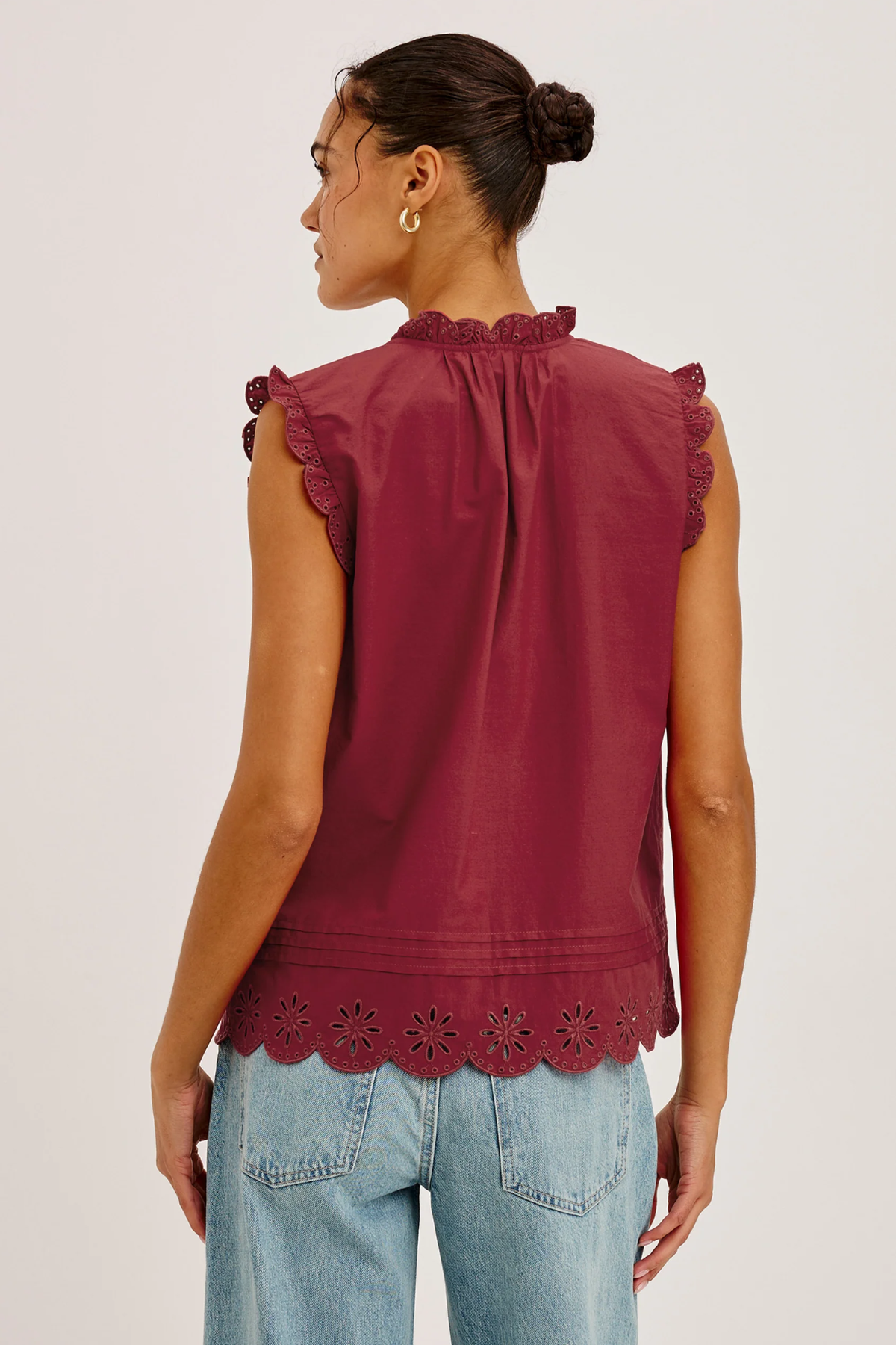 Woman wearing a burgundy sleeveless top with ruffle details and light blue jeans on a plain background