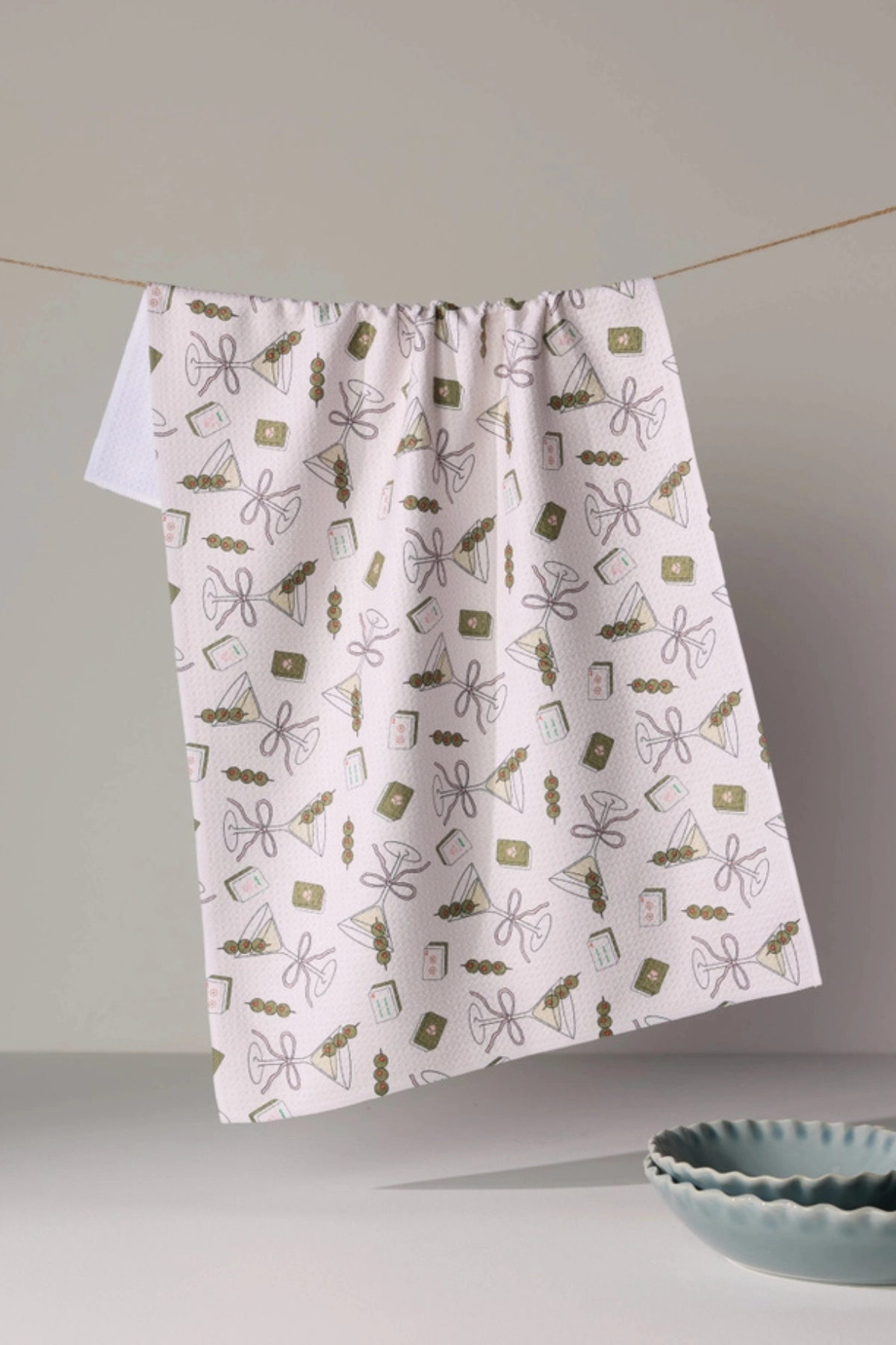 A white kitchen curtain with a delicate pattern of small brown, olive green, and gray line drawings of bows, flowers, and kitchen items, hanging from a string against a neutral gray wall.