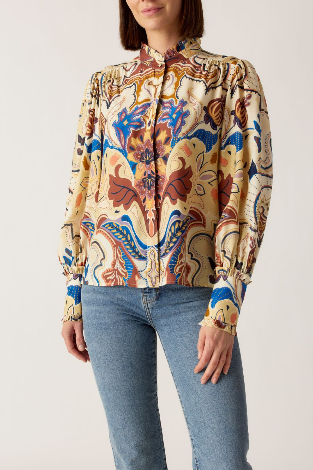 A woman wearing a long-sleeve high-neck blouse with a colorful abstract floral pattern in beige, blue, brown, and orange, paired with light-wash blue jeans against a plain light background.