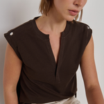 A woman wearing a dark brown sleeveless top with a split neckline and decorative silver buttons on the shoulders, paired with cream-colored pants, photographed against a neutral light gray background.