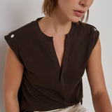 A woman wearing a dark brown sleeveless top with a split neckline and decorative silver buttons on the shoulders, paired with cream-colored pants, photographed against a neutral light gray background.