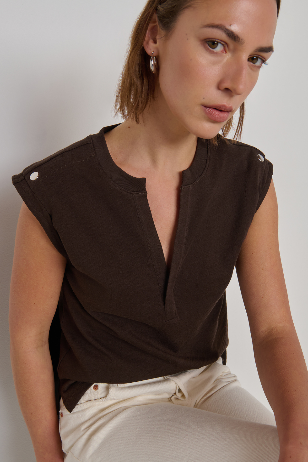 A woman wearing a dark brown sleeveless top with a split neckline and decorative silver buttons on the shoulders, paired with cream-colored pants, photographed against a neutral light gray background.