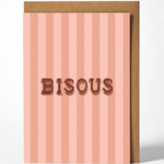 A greeting card with vertical pink and peach stripes, featuring the word 'BISOUS' in raised brown letters, shown with a brown kraft envelope behind it on a light gray background.