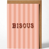 A greeting card with vertical pink and peach stripes, featuring the word 'BISOUS' in raised brown letters, shown with a brown kraft envelope behind it on a light gray background.