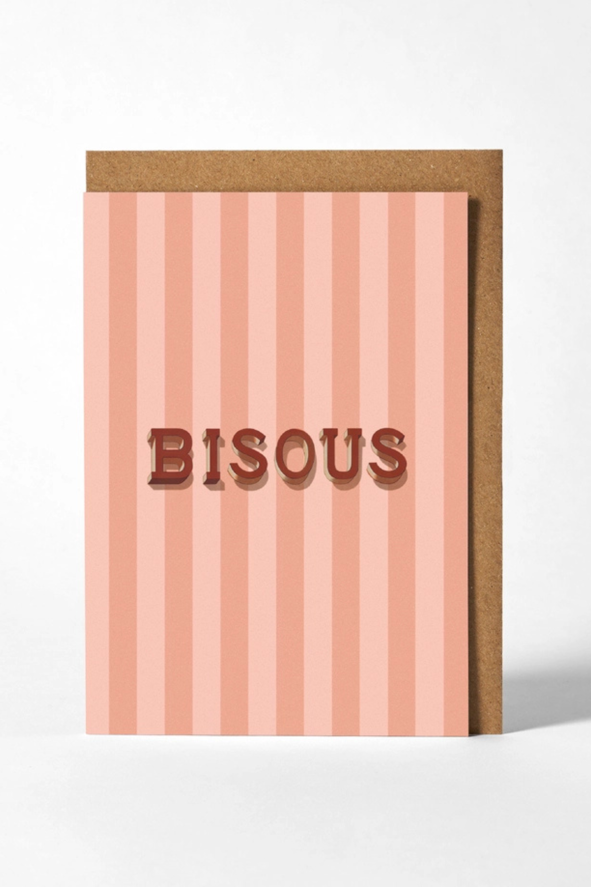 A greeting card with vertical pink and peach stripes, featuring the word 'BISOUS' in raised brown letters, shown with a brown kraft envelope behind it on a light gray background.