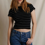 A woman modeling a black short-sleeve crop top with thin white horizontal stripes, paired with blue jeans, against a neutral beige fabric backdrop.