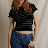 A woman modeling a black short-sleeve crop top with thin white horizontal stripes, paired with blue jeans, against a neutral beige fabric backdrop.
