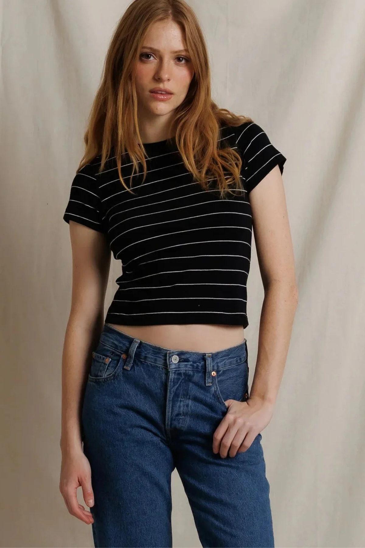 A woman modeling a black short-sleeve crop top with thin white horizontal stripes, paired with blue jeans, against a neutral beige fabric backdrop.