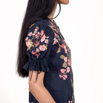 A woman shown in profile wearing a navy blue short-sleeve blouse with pink and green floral embroidery and tie details on the sleeve cuffs, against a plain white background.