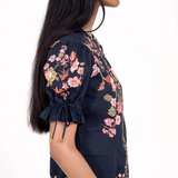 A woman shown in profile wearing a navy blue short-sleeve blouse with pink and green floral embroidery and tie details on the sleeve cuffs, against a plain white background.