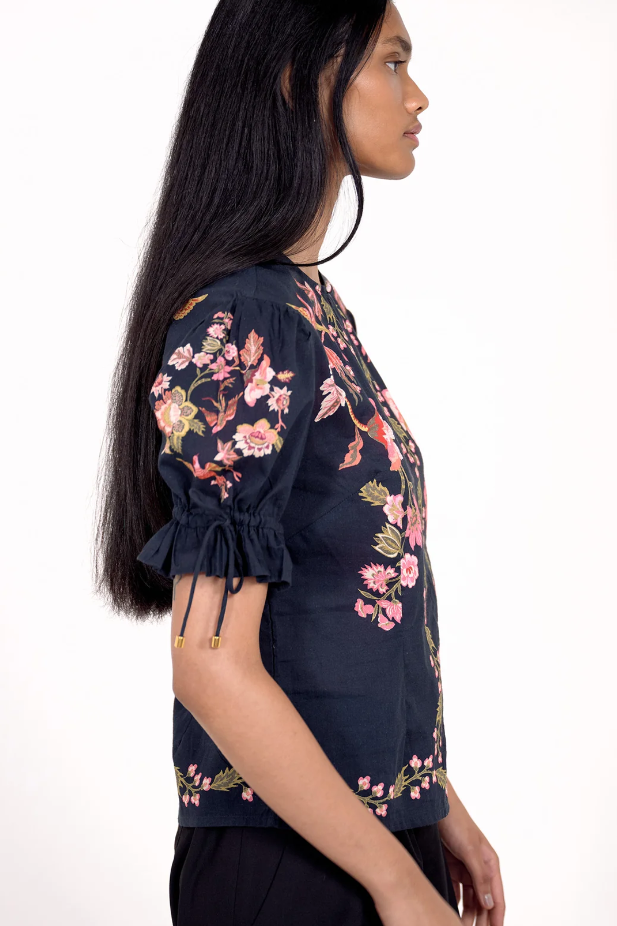 A woman shown in profile wearing a navy blue short-sleeve blouse with pink and green floral embroidery and tie details on the sleeve cuffs, against a plain white background.