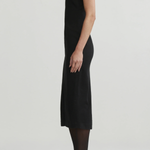 A model shown in profile wearing a sleeveless black midi dress, sheer black tights, and black pointed-toe heels against a plain light gray studio background.