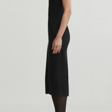 A model shown in profile wearing a sleeveless black midi dress, sheer black tights, and black pointed-toe heels against a plain light gray studio background.
