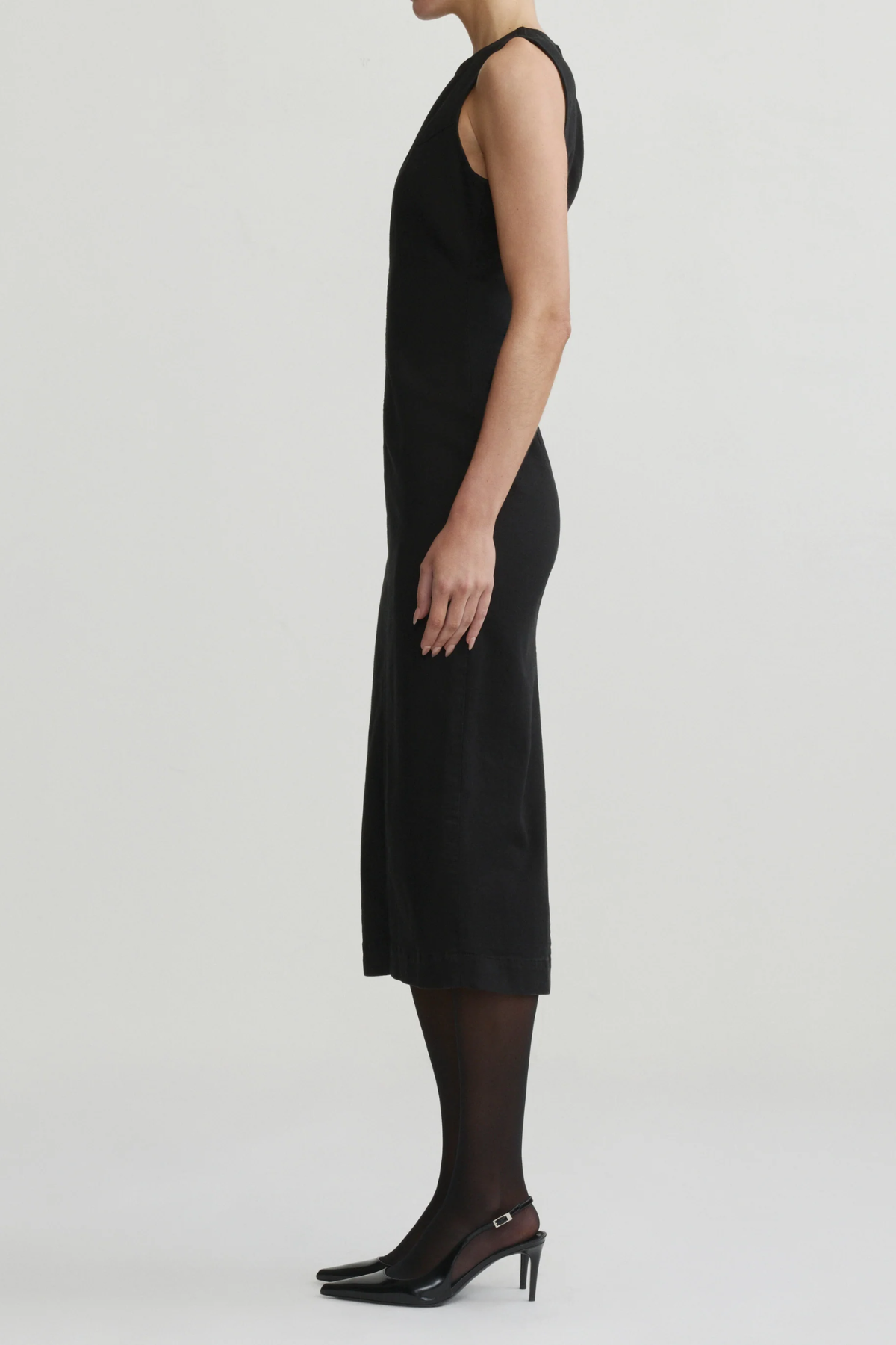 A model shown in profile wearing a sleeveless black midi dress, sheer black tights, and black pointed-toe heels against a plain light gray studio background.