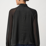 A woman shown from the back wearing a black sheer long-sleeve blouse with opaque raglan sleeves and dark patterned pants, against a light gray studio background.