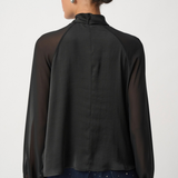 A woman shown from the back wearing a black sheer long-sleeve blouse with opaque raglan sleeves and dark patterned pants, against a light gray studio background.