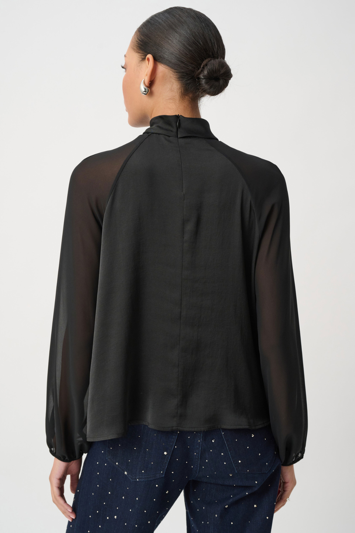A woman shown from the back wearing a black sheer long-sleeve blouse with opaque raglan sleeves and dark patterned pants, against a light gray studio background.