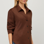 A woman wearing a brown button-front collared shirt with three-quarter sleeves and blue jeans, photographed against a plain light gray studio background.