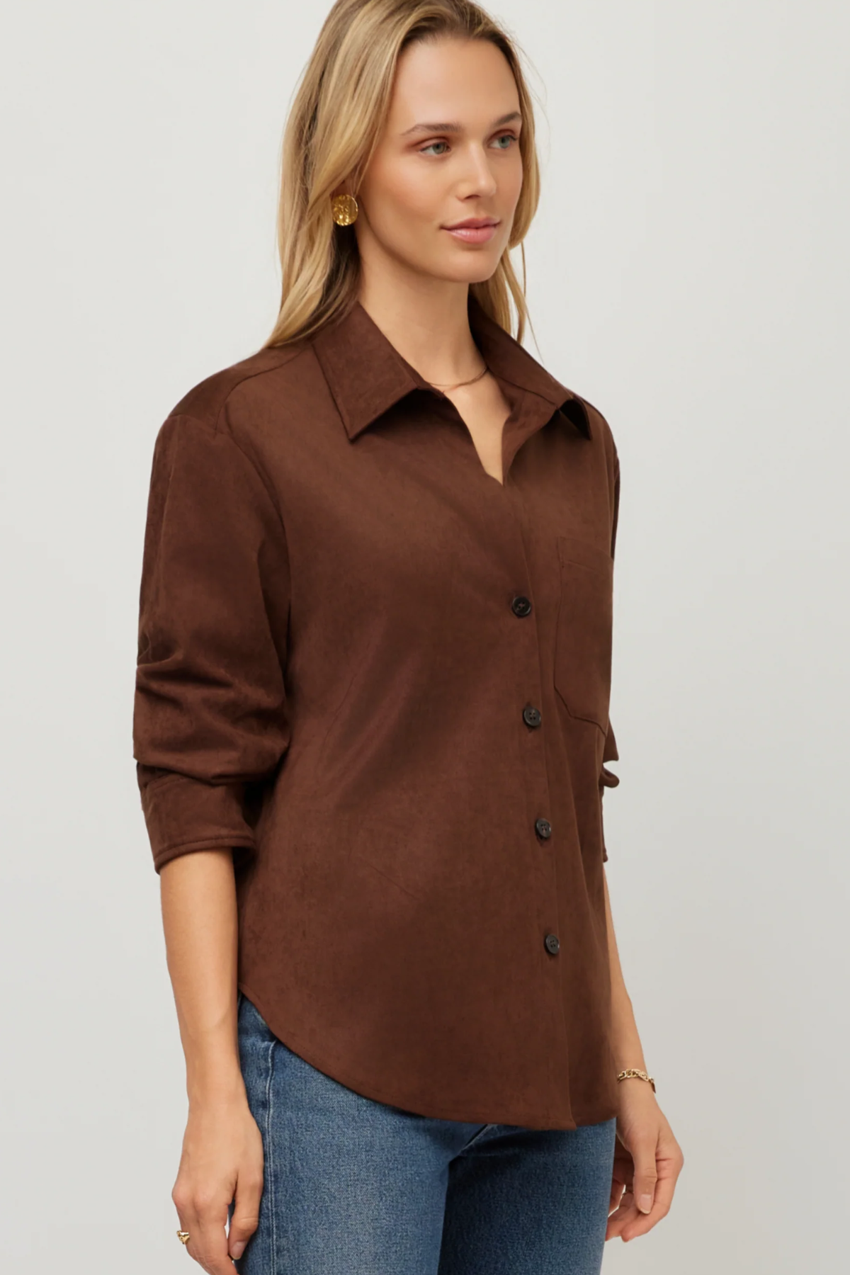 A woman wearing a brown button-front collared shirt with three-quarter sleeves and blue jeans, photographed against a plain light gray studio background.