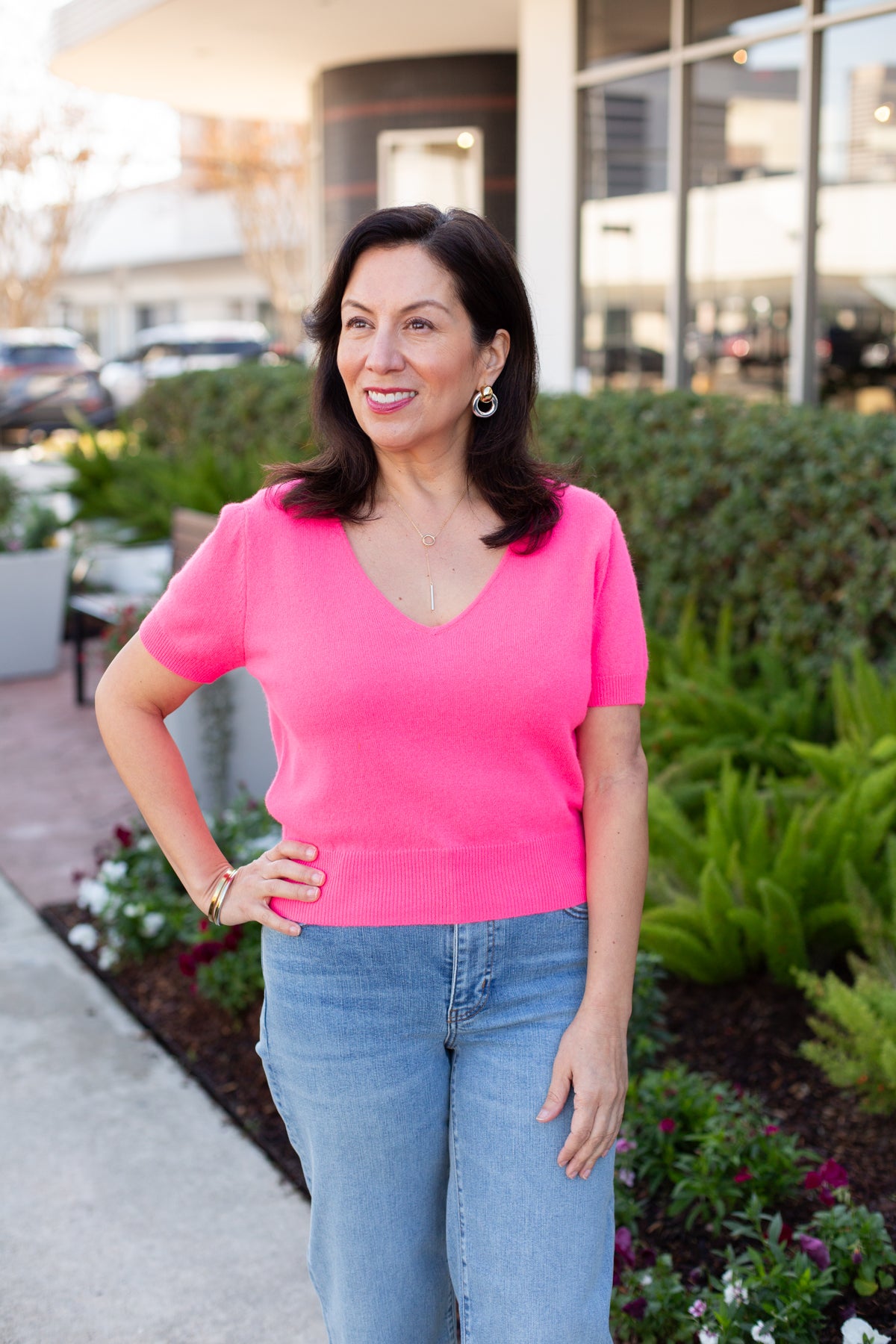 Woman in a pink shirt and blue jeans standing outdoors with greenery and buildings in the background