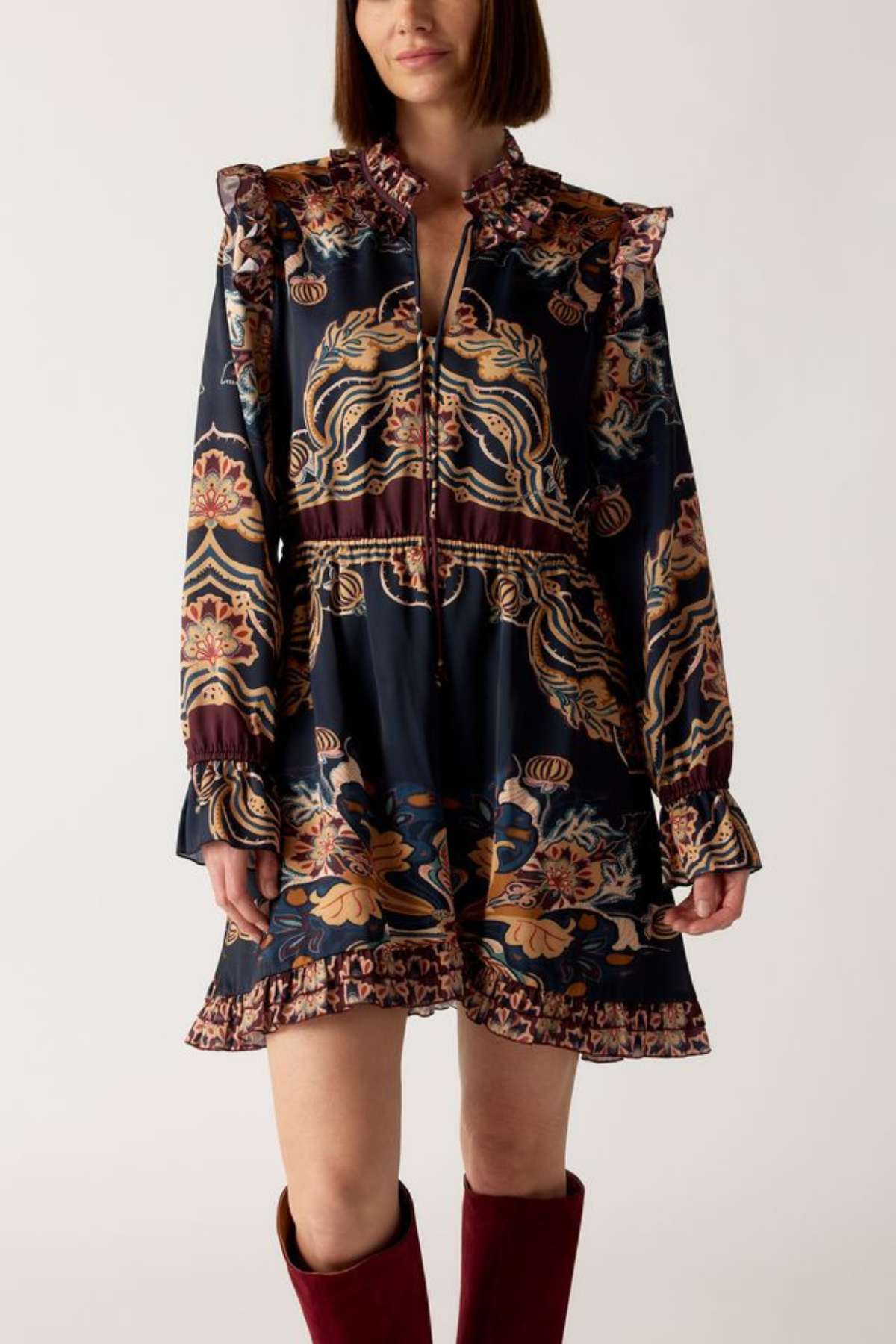 A model wearing a dark navy blue mini dress with a complex gold, brown, and burgundy bohemian-style print, paired with burgundy knee-high boots, against a plain light gray background.
