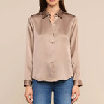 A woman modeling a taupe satin button-down shirt with blue jeans, standing against a solid beige background.