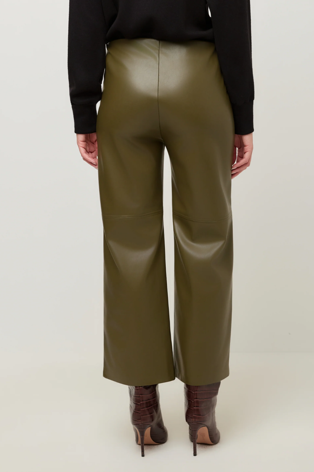 A person shown from the back wearing olive green wide-leg faux leather pants, a black long-sleeve top, and brown heeled ankle boots against a plain light background.