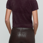 Rear view of a model wearing a short-sleeve knit polo shirt in deep plum purple and dark brown faux-leather pants, against a light gray studio background.
