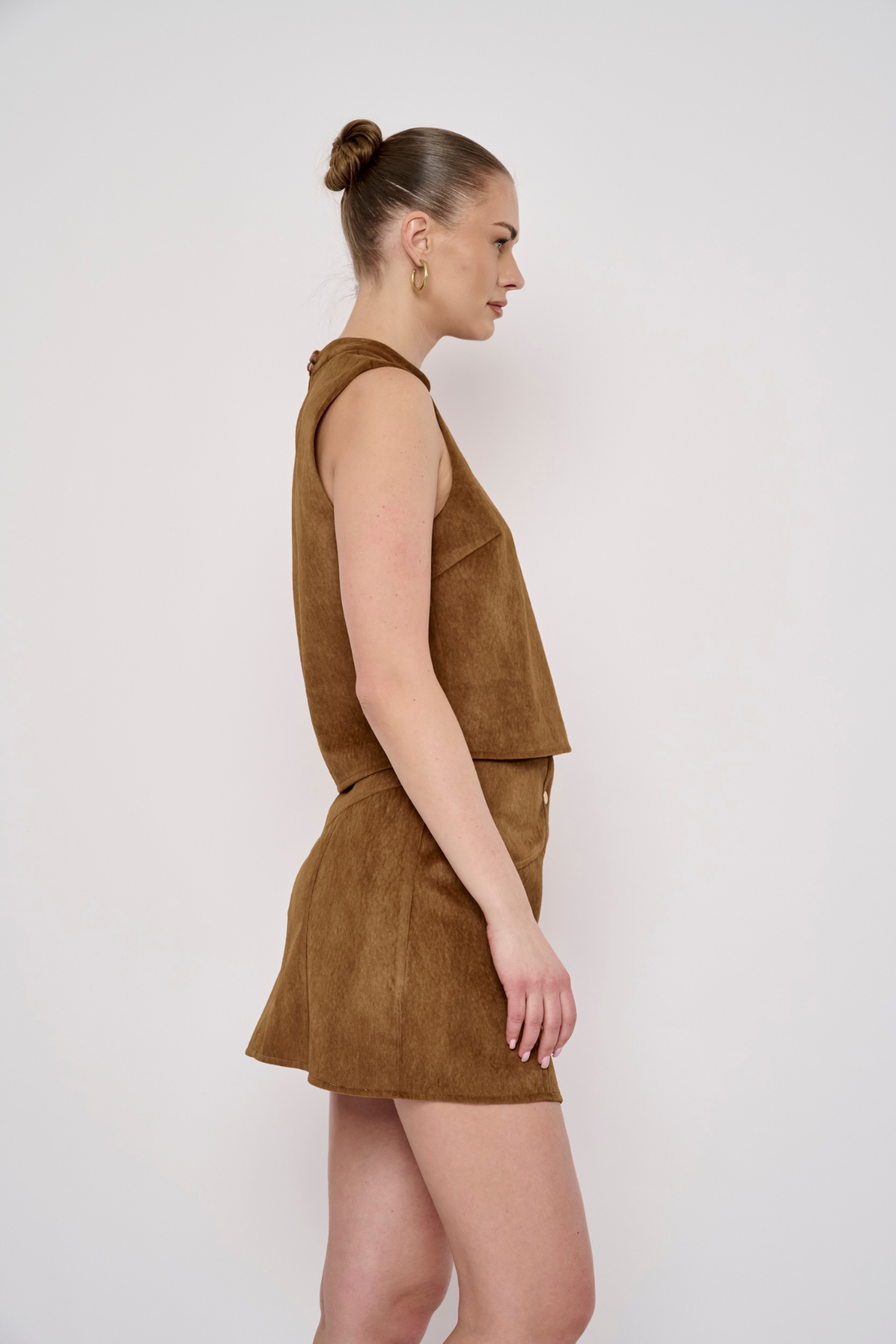 A woman in side profile wearing a sleeveless brown faux-suede romper with a matching mini skirt, set against a plain light gray studio background.
