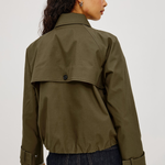 A woman shown from the back wearing an olive green utility jacket with a buttoned flap detail and dark blue jeans, set against a plain light gray studio background.
