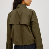 A woman shown from the back wearing an olive green utility jacket with a buttoned flap detail and dark blue jeans, set against a plain light gray studio background.