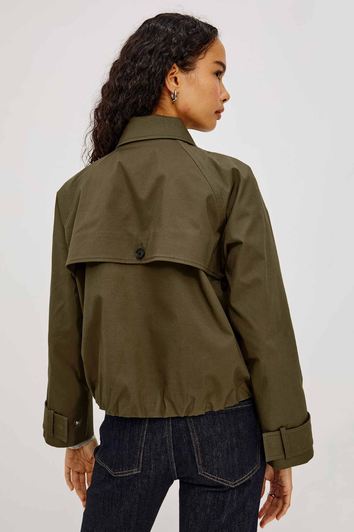 A woman shown from the back wearing an olive green utility jacket with a buttoned flap detail and dark blue jeans, set against a plain light gray studio background.