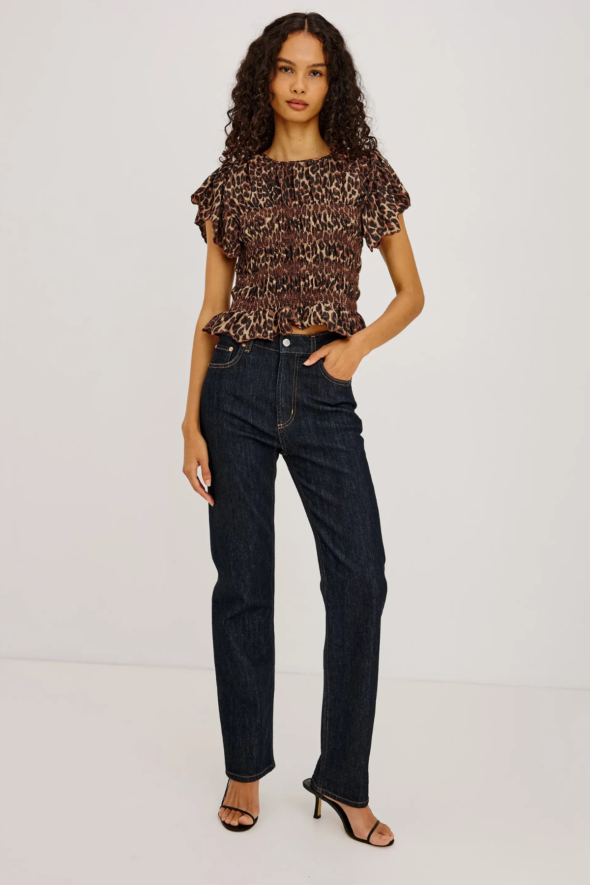 A model wearing a brown and black leopard-print ruffled short-sleeve top, dark wash blue jeans, and black heeled sandals, standing against a plain white studio background.