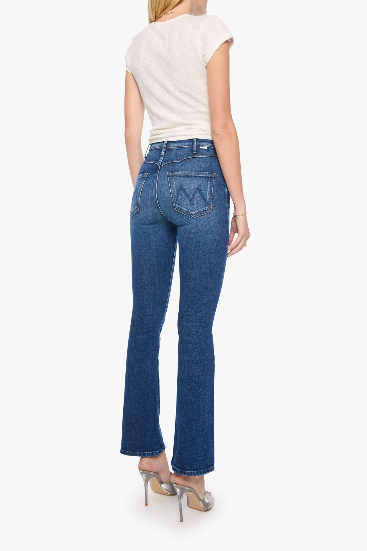 A woman shown from the back wearing blue high-rise flare jeans, a cream short-sleeve top, and metallic high heels against a plain white background.