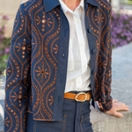 A person wearing a navy blue button-front jacket with orange geometric and floral patterns, layered over a white collared shirt and paired with dark blue jeans and a tan belt, photographed outdoors against a blurred stone and plant background.