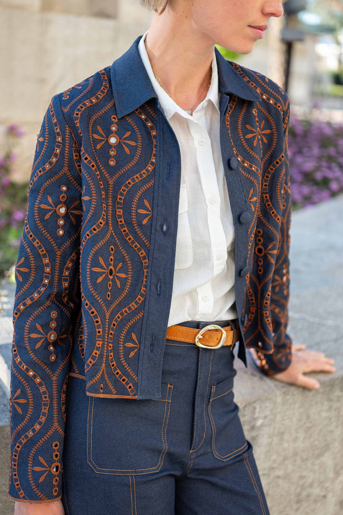 A person wearing a navy blue button-front jacket with orange geometric and floral patterns, layered over a white collared shirt and paired with dark blue jeans and a tan belt, photographed outdoors against a blurred stone and plant background.
