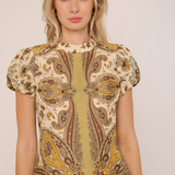A woman wearing a short-sleeve top with a beige, brown, and yellow paisley pattern, photographed against a plain light background.