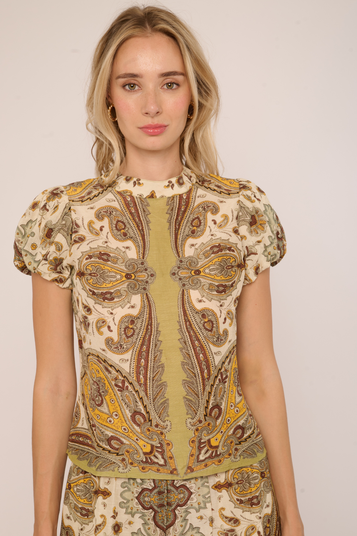 A woman wearing a short-sleeve top with a beige, brown, and yellow paisley pattern, photographed against a plain light background.