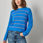 A woman wearing a bright blue knit sweater with thin pink horizontal stripes and blue jeans, standing against a light gray studio background.