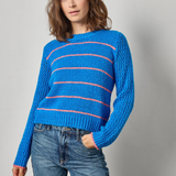 A woman wearing a bright blue knit sweater with thin pink horizontal stripes and blue jeans, standing against a light gray studio background.