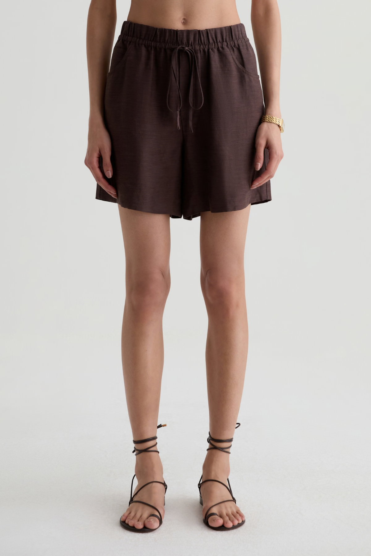 A model wearing dark brown drawstring shorts and matching strappy flat sandals, shown from the waist down against a light gray studio background.