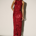 A woman modeling a sleeveless, floor-length red dress with a tonal floral pattern and high neckline, standing against a plain off-white wall.