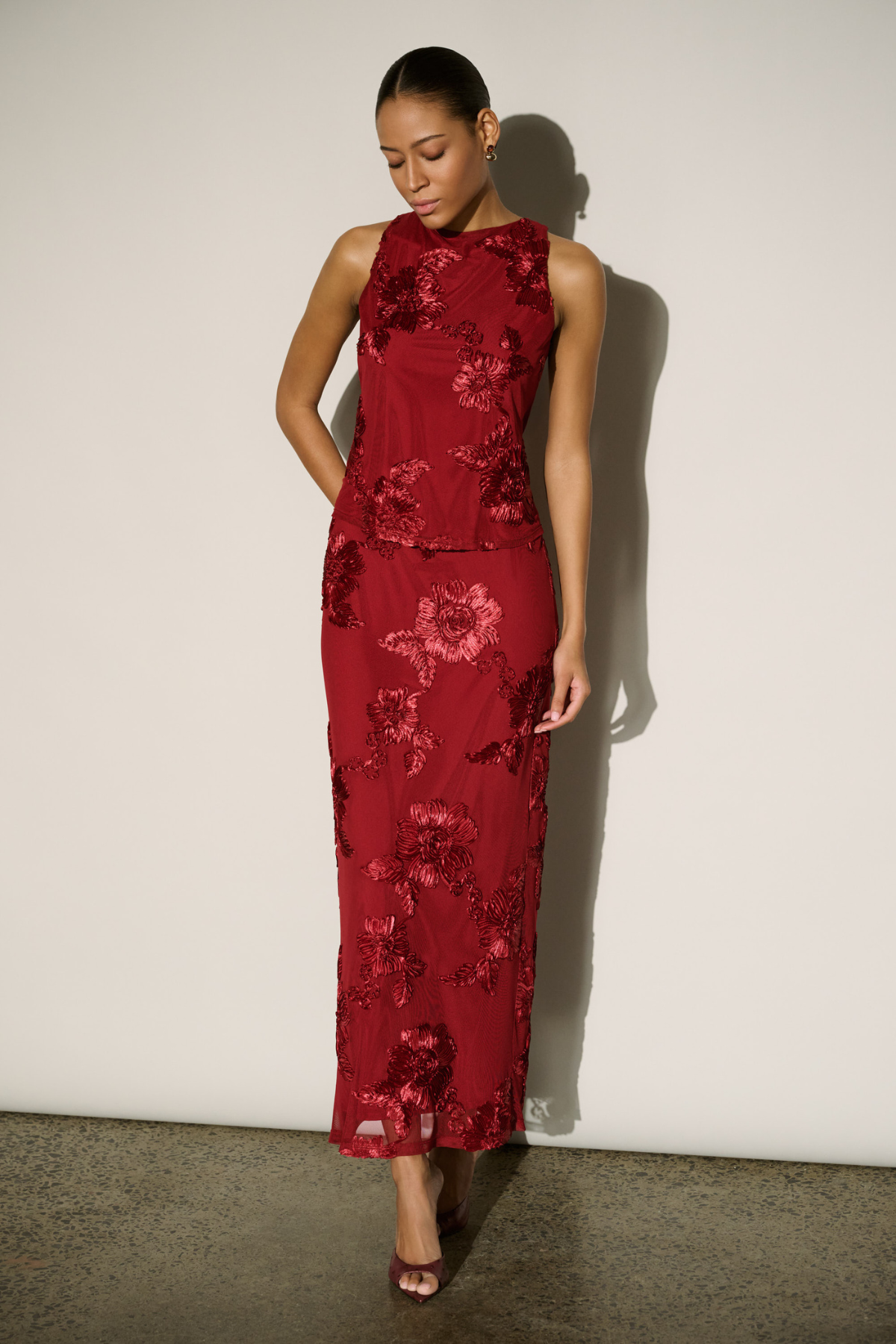 A woman modeling a sleeveless, floor-length red dress with a tonal floral pattern and high neckline, standing against a plain off-white wall.