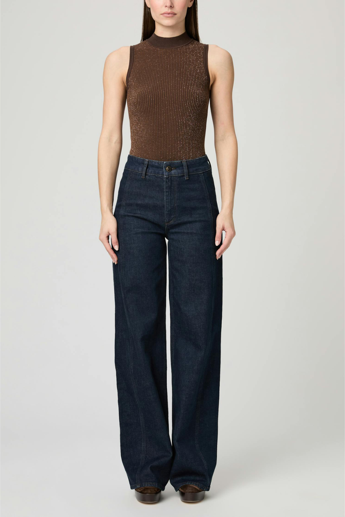 A model wearing a brown ribbed sleeveless mock-neck top and dark blue wide-leg jeans, standing against a light gray studio background.