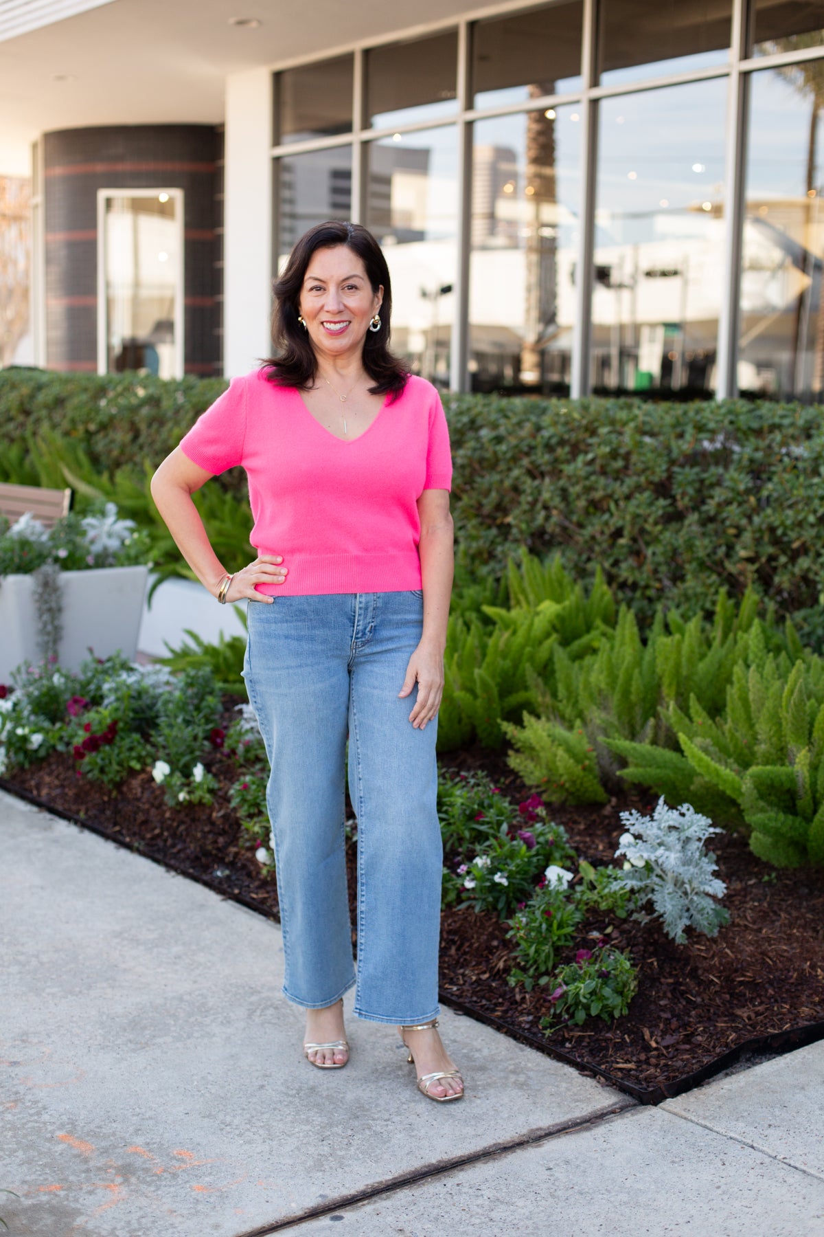 Woman in a pink shirt and blue jeans standing outdoors near a building with greenery.