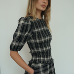 A woman wearing a black, white, and gray plaid dress with puffed short sleeves and a smocked waist, standing against a plain light gray wall.