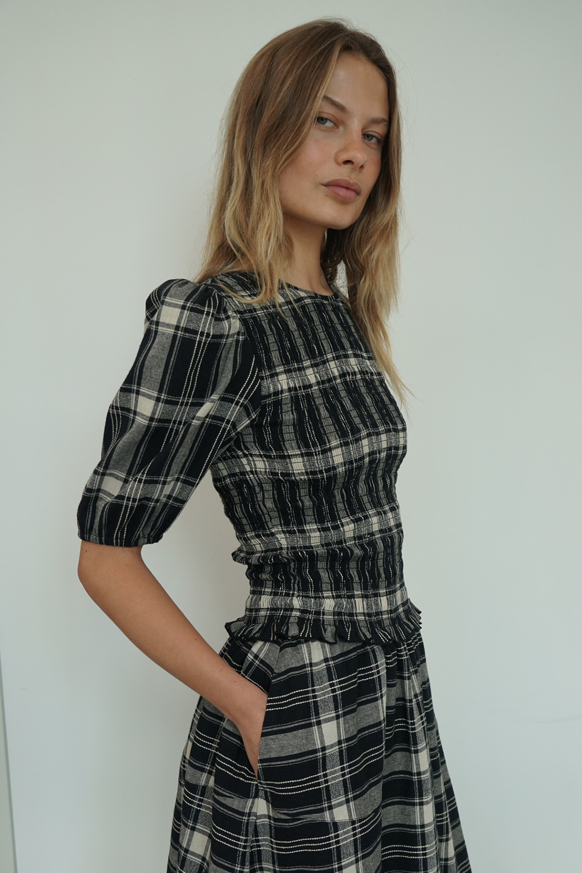 A woman wearing a black, white, and gray plaid dress with puffed short sleeves and a smocked waist, standing against a plain light gray wall.
