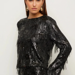 A woman modeling a black long-sleeve sequined top with fringe detailing, paired with a matching black fringed skirt, against a plain light background.