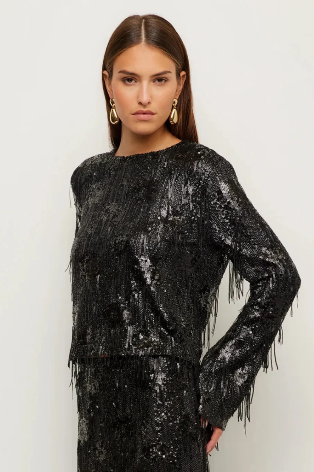 A woman modeling a black long-sleeve sequined top with fringe detailing, paired with a matching black fringed skirt, against a plain light background.