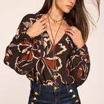 A woman wearing a brown long-sleeve blouse with a beige, black, and rust-red abstract pattern, paired with dark blue jeans featuring gold button details on the pocket, set against a plain beige background.